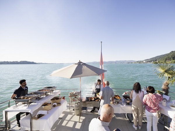 Aussendeck mit Gästen am BBQ Buffet mit Sicht über den See im Hintergrund